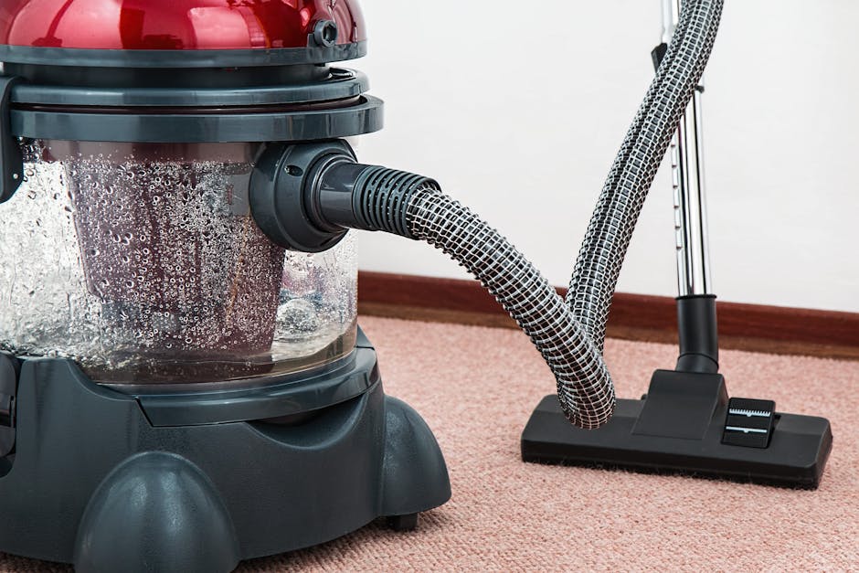 A woman is vacuuming a patterned area rug with a yellow vacuum cleaner in a living room. The room has wooden flooring surrounding the rug, a red leather sofa visible on the right, and natural light il